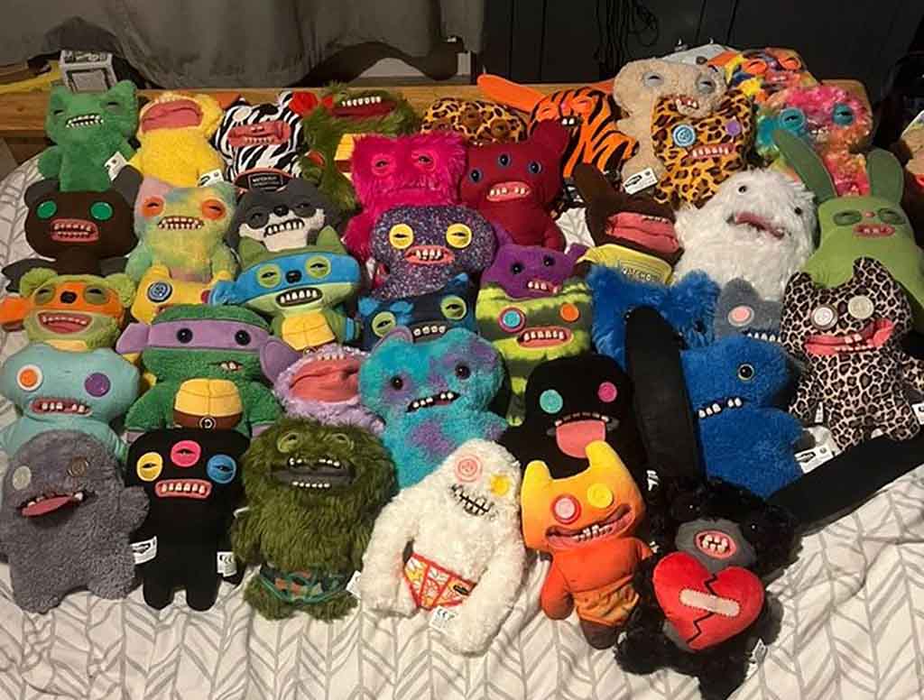 A large assorted collection of Fuggler plush toys displayed on a bed, featuring many colors, patterns, and facial expressions with their signature goofy teeth and button eyes.