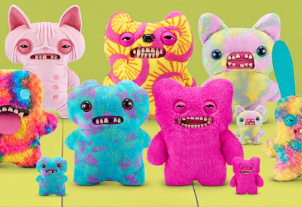 A vibrant assortment of Fuggler plush toys in various neon and pastel colors, each featuring quirky faces, toothy grins, button eyes, and unique monster designs, displayed together against a bright green background.