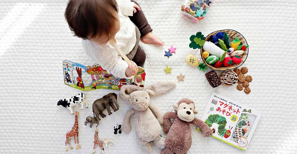 A baby sits on a soft white mat surrounded by plush toys, animal figurines, books, and colorful learning materials while playing and exploring.