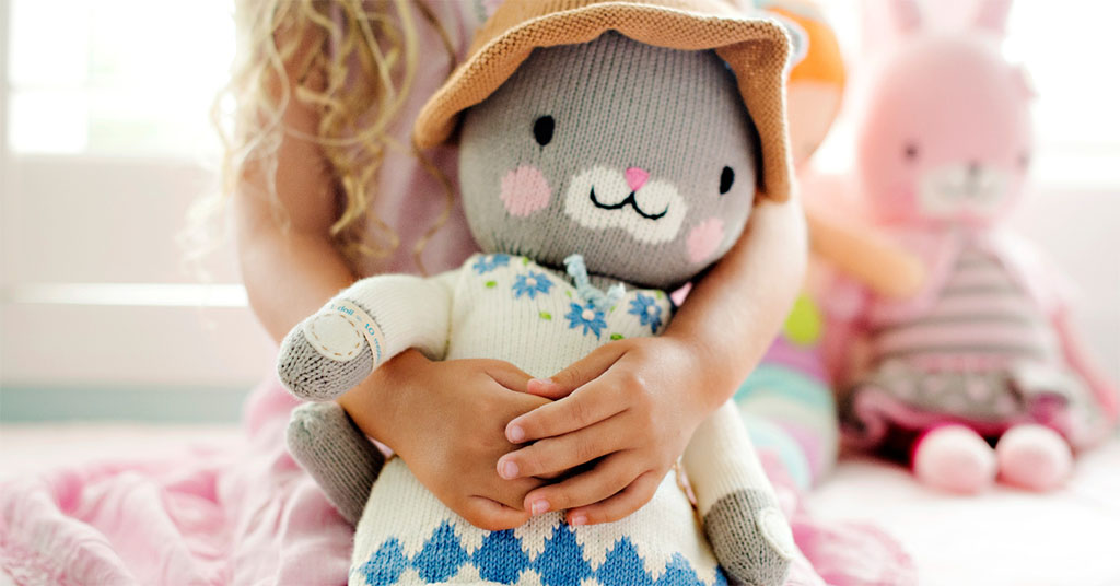Child hugging a knitted bunny doll with other plush toys in the background.