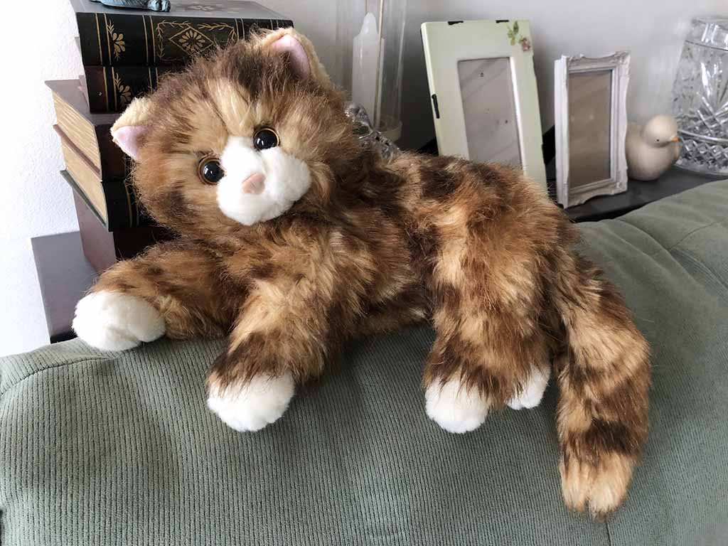 A realistic brown tabby cat plush toy with long fur, white paws, and large expressive eyes, posed on a green sofa with home décor elements in the background.