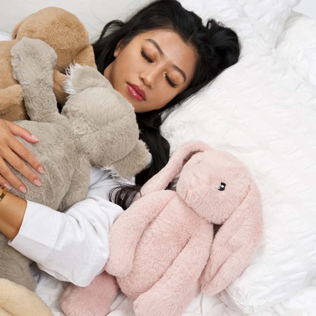 A woman peacefully sleeps on a white bed while cuddling several soft, long-eared plush bunny toys, illustrating the comfort and emotional support stuffed animals provide for adults.