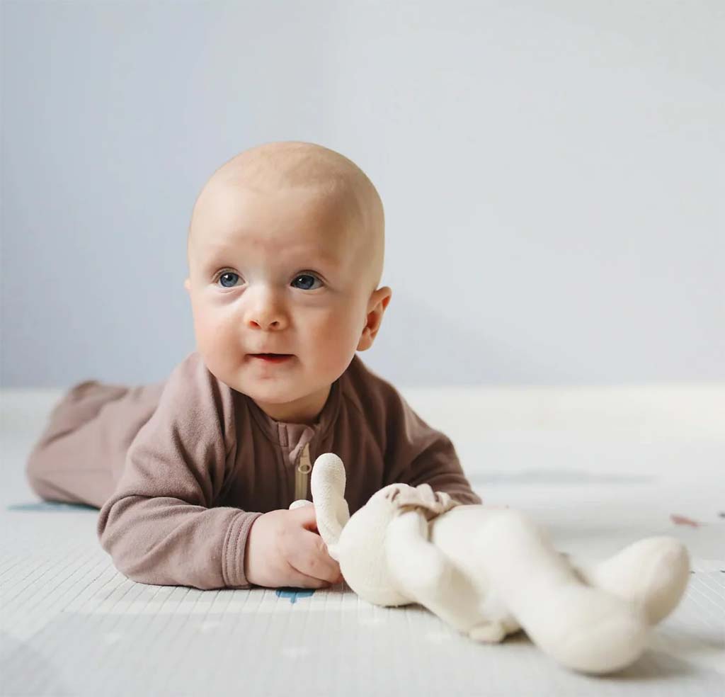 A baby lying on their tummy on a soft play mat while gently holding a cream-colored plush toy, looking up with a calm expression.
