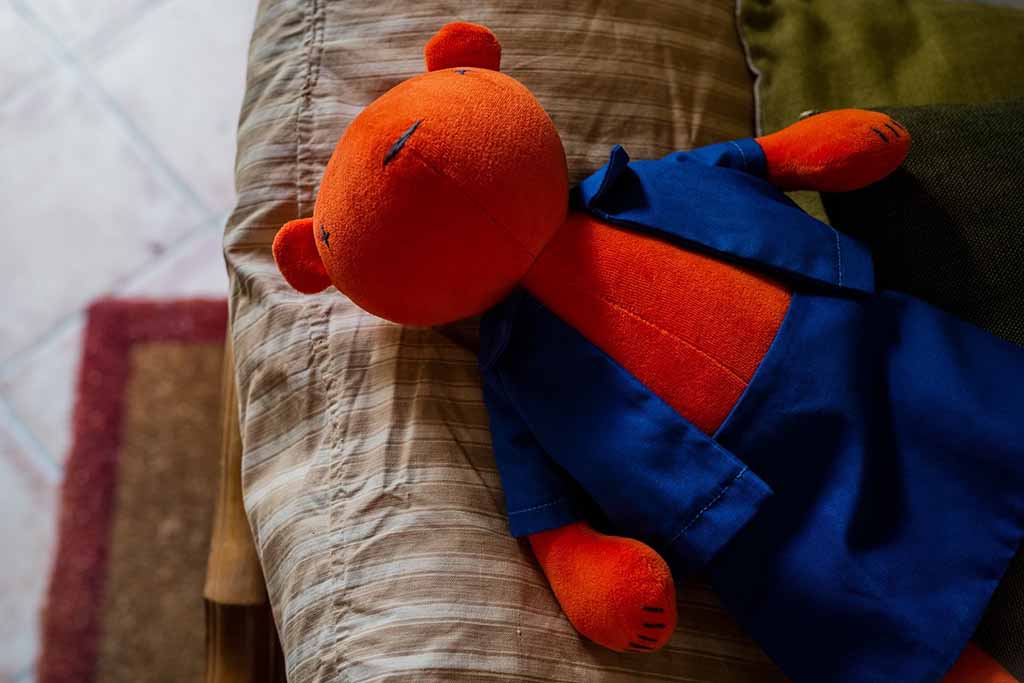 Orange stuffed bear wearing blue overalls lying on a sofa cushion, softly lit in a cozy home setting.