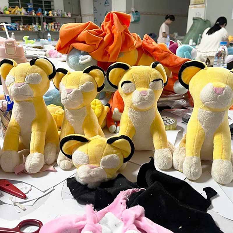 Several yellow lion plush toys in various stages of assembly on a cluttered workshop table with fabric pieces and tools.