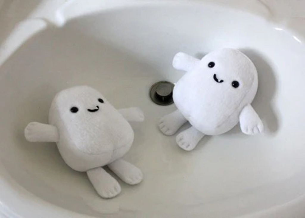 Two small white plush toys with smiling faces floating in a bathroom sink filled with water, viewed from above.