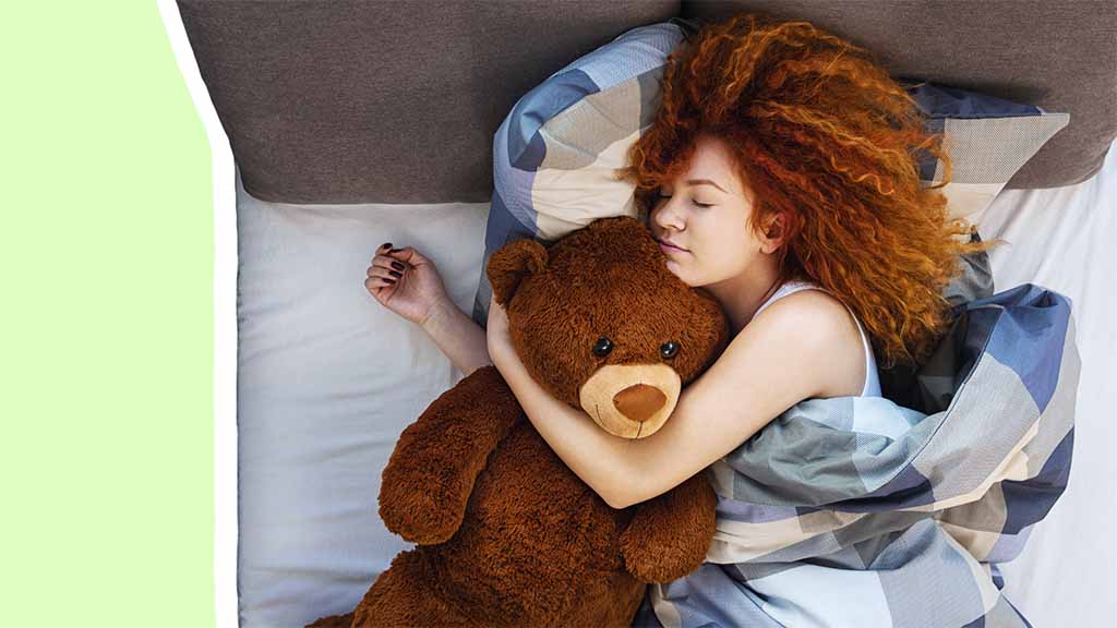 A young woman with curly red hair sleeps in bed while hugging a large brown teddy bear, showing how plush toys provide comfort and emotional support for adults.