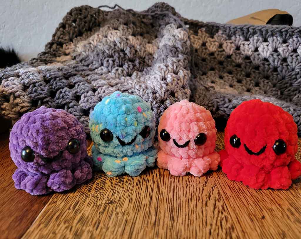 Four small, round, chenille-textured octopus plushies in purple, blue, pink, and red, each with shiny black eyes and a cute smiling face, sitting on a wooden surface in front of a gray crocheted blanket.