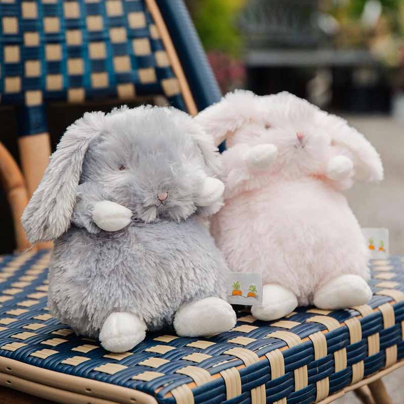 Two soft bunny plush toys sitting on a woven chair, one gray and one pink.