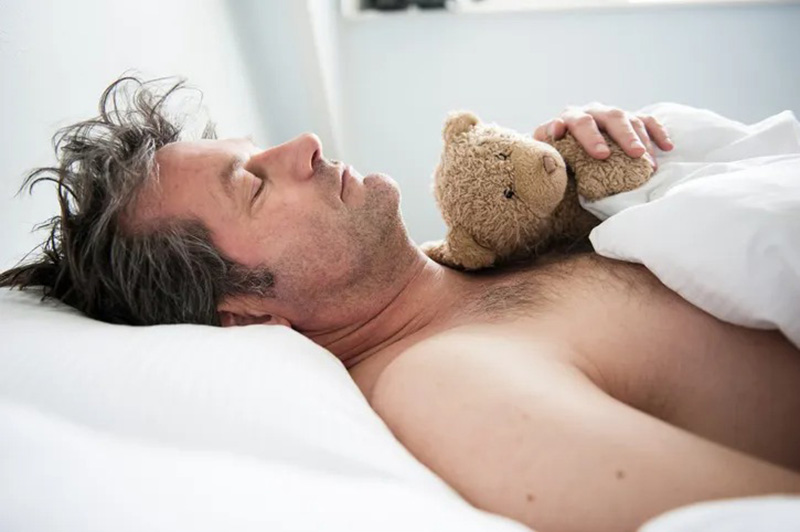 An adult man sleeps peacefully in bed while holding a small teddy bear against his chest, showing the comfort and emotional support plush toys can provide for grown-ups.