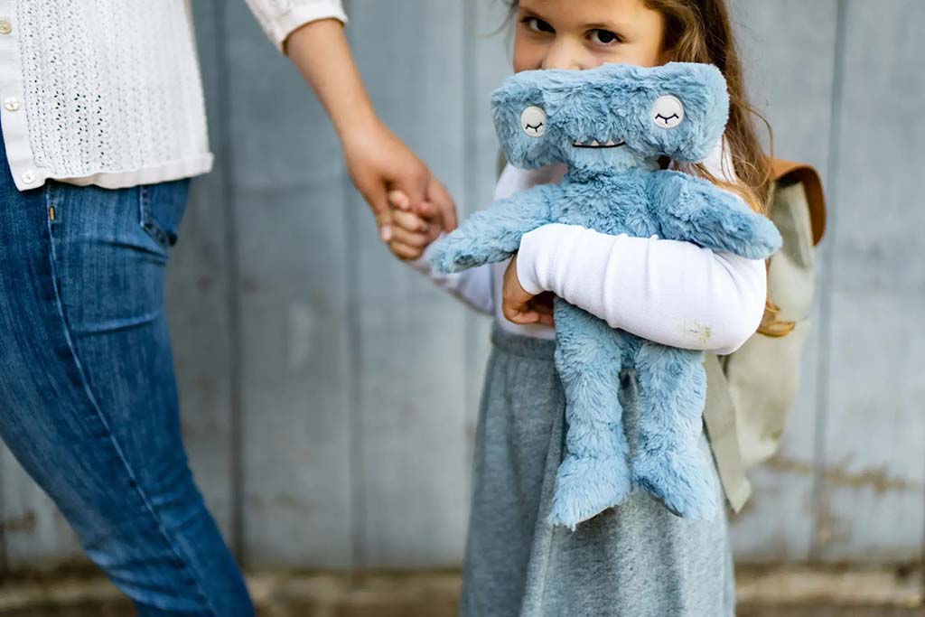 Young child holding a soft blue plush toy while walking hand in hand with an adult, outdoors against a gray wall.