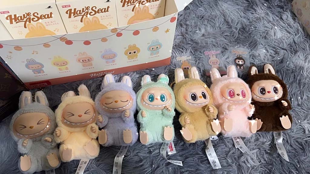Row of pastel-colored big-eyed bunny plush keychain dolls from a blind box series, each with vinyl face details, displayed on a soft gray surface with branded packaging behind.