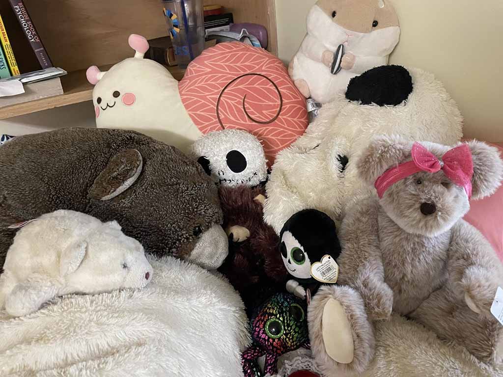 A cozy pile of assorted plush toys and stuffed animals arranged on a shelf, including bears, rabbits, character plushies, and soft cushions in neutral and pastel colors.