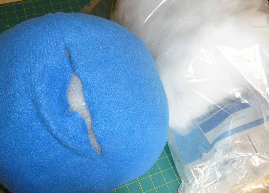 Blue plush cushion with a visible tear exposing white foam filling inside, placed next to a clear plastic bag filled with stuffing material on a cutting mat, illustrating damaged plush toy filling.