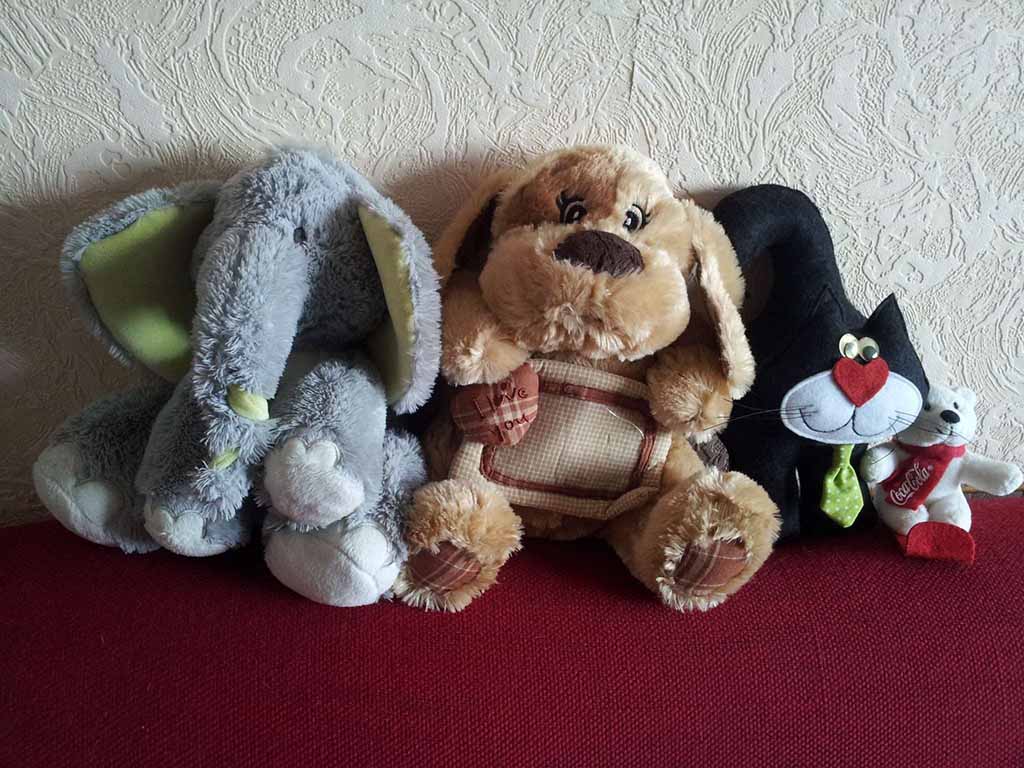 Four stuffed animals sitting side by side on a red sofa, including a gray elephant plush, a brown dog plush, a black cat plush with a green tie, and a small white bear plush, arranged against a textured wall.