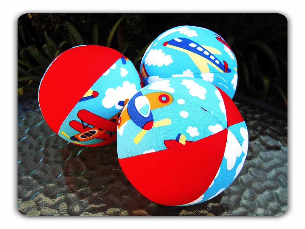 Three handmade fabric plush balls with red panels and blue airplane-and-cloud print, arranged on a glass tabletop, showing soft textile texture and stitched seams.