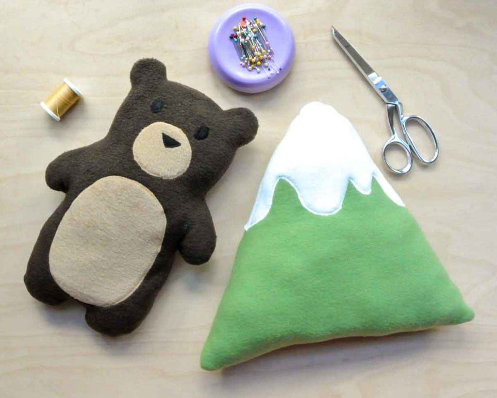 A handmade felt bear plush and a mountain-shaped plush placed on a table with sewing tools, pins, thread, and scissors.