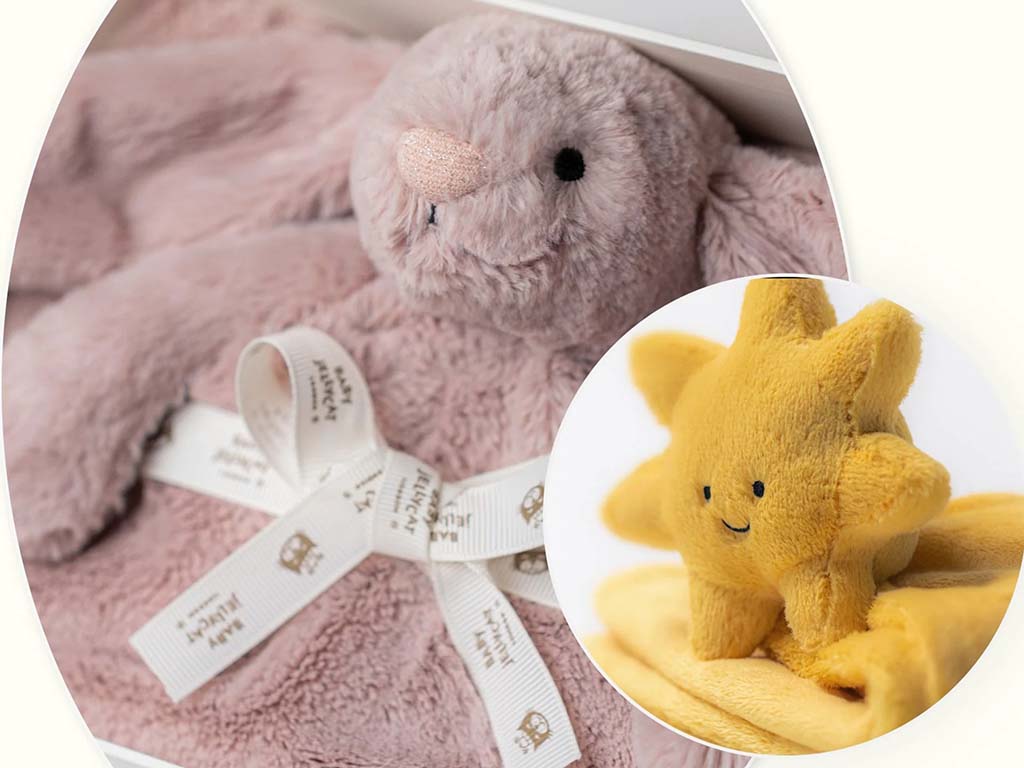 Close-up of a soft pink Jellycat bunny comforter with branded ribbon, paired with a small yellow sun plush toy on a matching blanket, styled for a baby gift set.