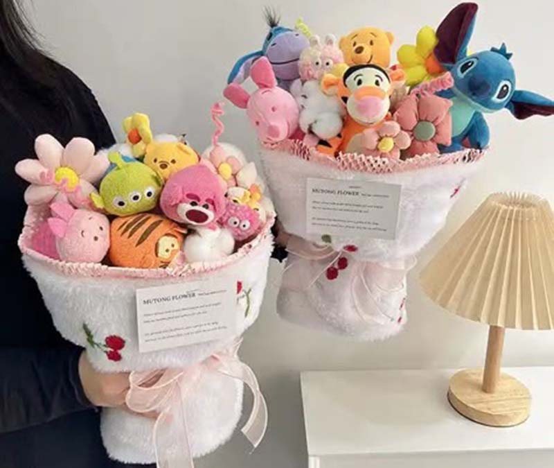 Two large plush toy bouquets filled with colorful character plushies, wrapped in soft white fabric with embroidered flowers and pink lace ribbons, held beside a small table lamp.