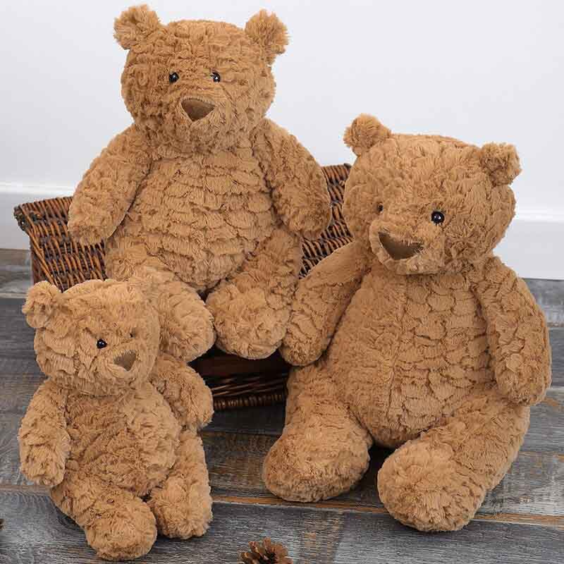 Three soft brown teddy bears in small, medium, and large sizes sitting together on a wooden floor beside a wicker basket, showcasing plush toys with textured fur ideal for cuddling and gifting.