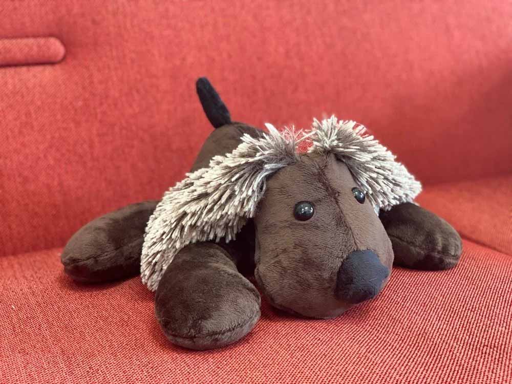 A soft brown plush dog with floppy legs, shaggy light-brown ears, and a black nose lying on a red fabric sofa.