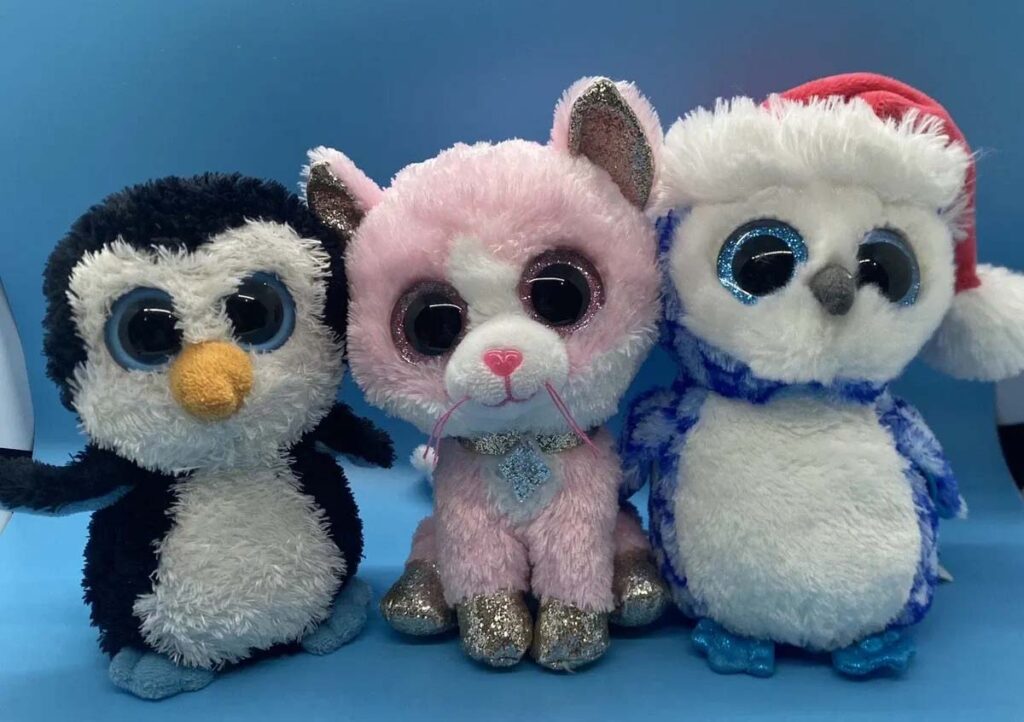 Three cute plush toys—a penguin, a pink cat, and an owl with a Santa hat—featuring oversized glittery eyes