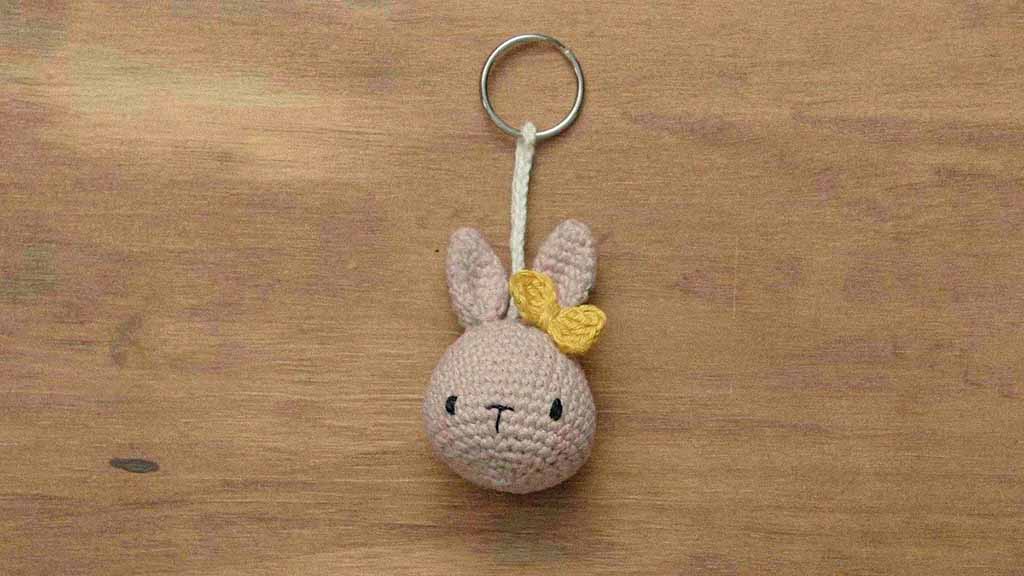 Small handmade crochet bunny keychain with beige yarn, upright ears, simple stitched face, and a yellow bow, displayed on a wooden surface.