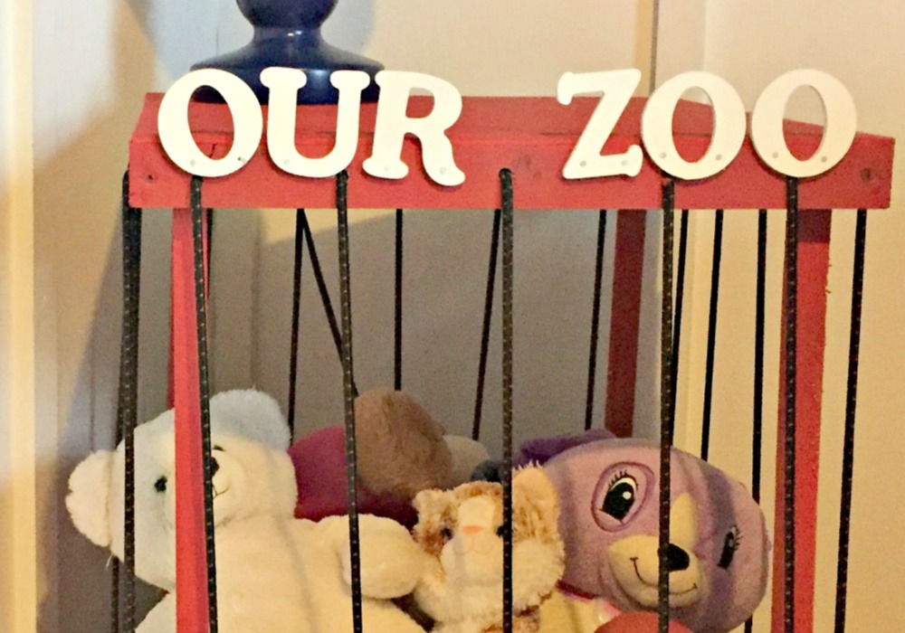 DIY stuffed animal zoo made from a red wooden frame with black bars, displaying plush toys inside and a white “Our Zoo” sign on top.