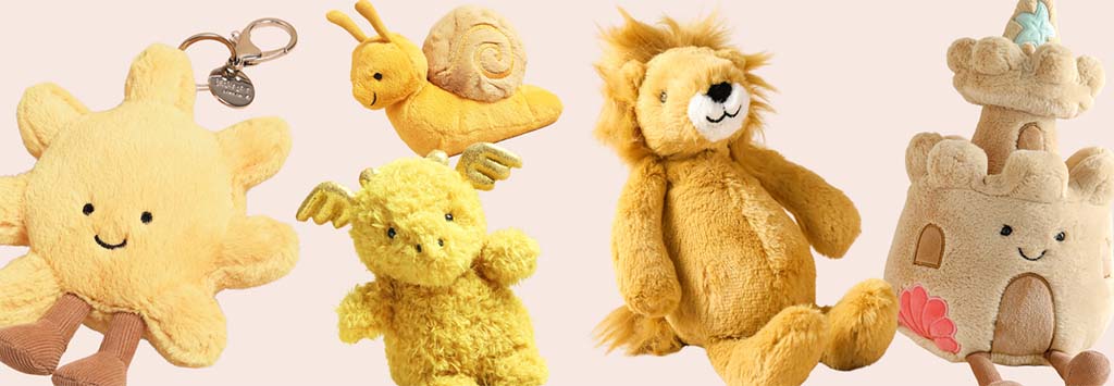 Assorted yellow Jellycat plush toys including a smiling sun, snail, dragon, lion, and sandcastle displayed together on a soft pastel background.