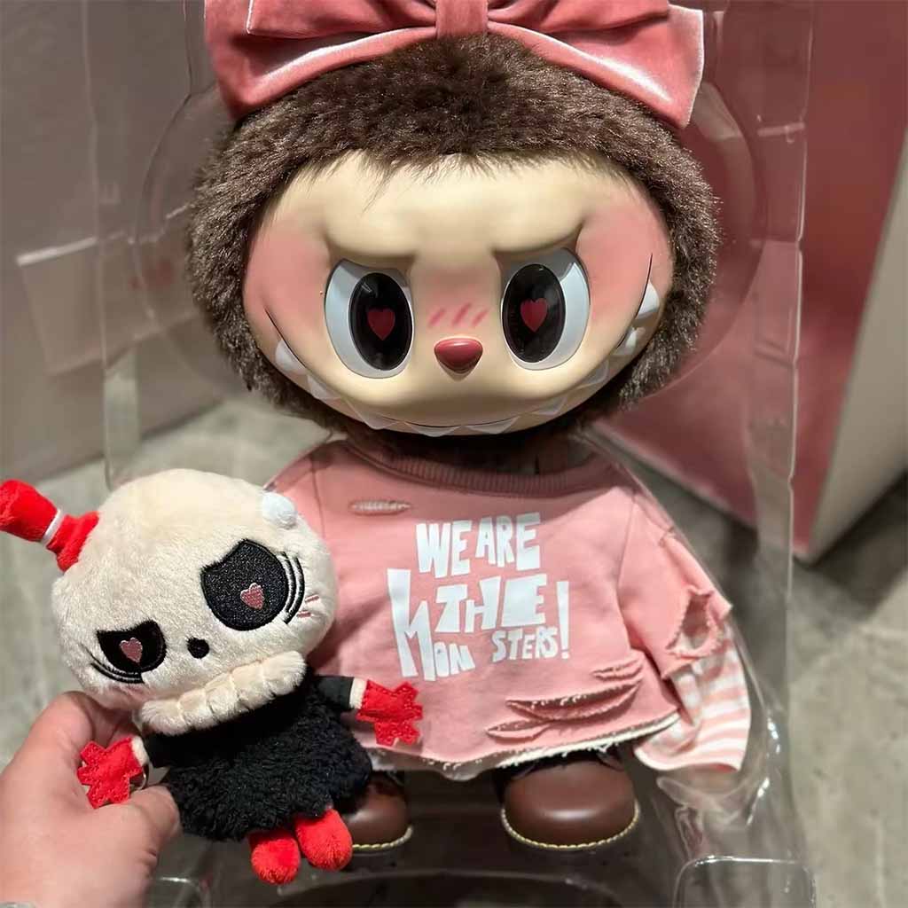 Big-eyed designer plush character with heart-shaped pupils, wearing a pink bow and printed outfit, displayed in clear packaging next to a small matching monster plush.