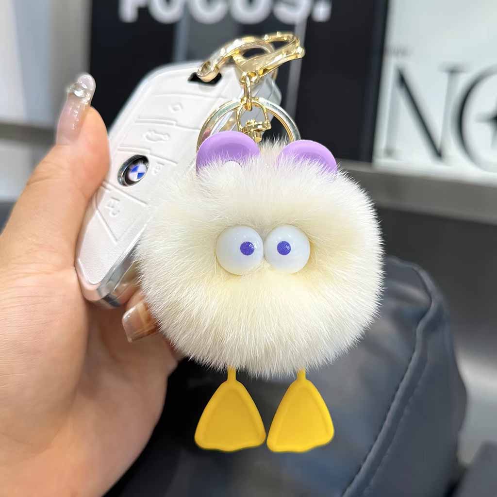 Cute fluffy white pom-pom keychain with big googly eyes, purple ear accents, and yellow feet, attached to a gold keyring held beside a car key.
