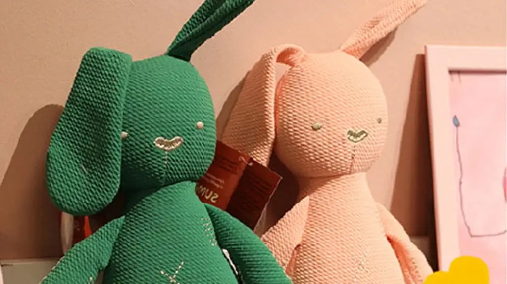 Two knitted plush bunny toys—one green and one light peach—are leaning against a wall. Both feature long ears, simple embroidered facial details, and a soft textured fabric. A framed drawing and decorative items appear in the background, creating a cozy display setting.