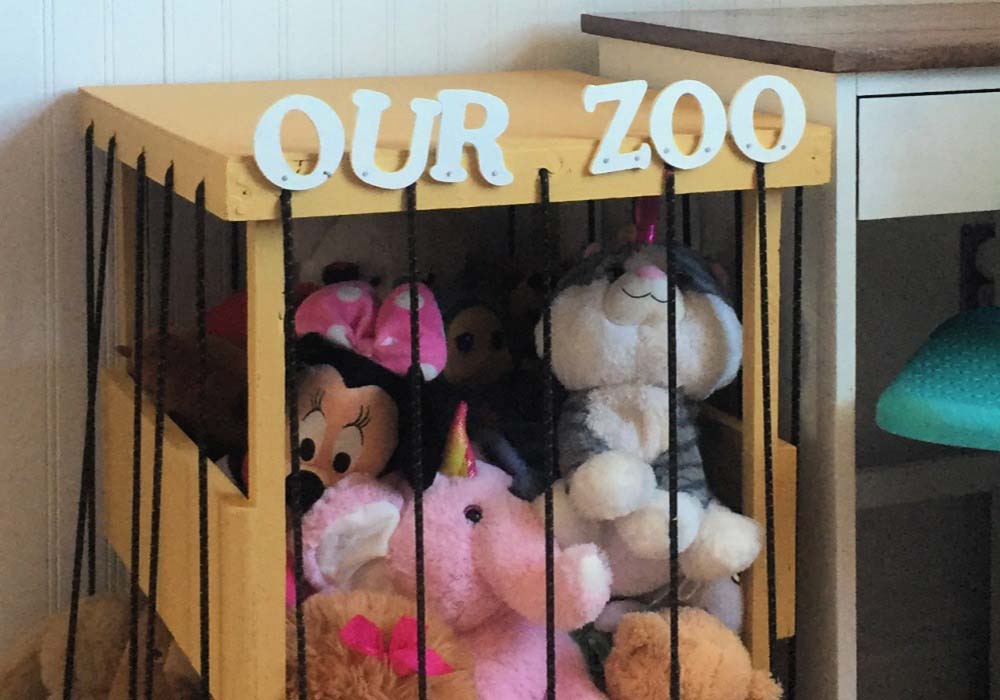 Wooden DIY stuffed animal zoo storage with black rope bars, labeled “Our Zoo,” holding plush toys like a unicorn, bear, and character dolls inside.