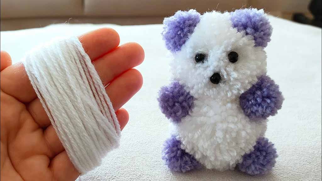 Hand holding white yarn beside a small handmade pom-pom panda plush featuring white body fluff with purple ears, arms, and feet.