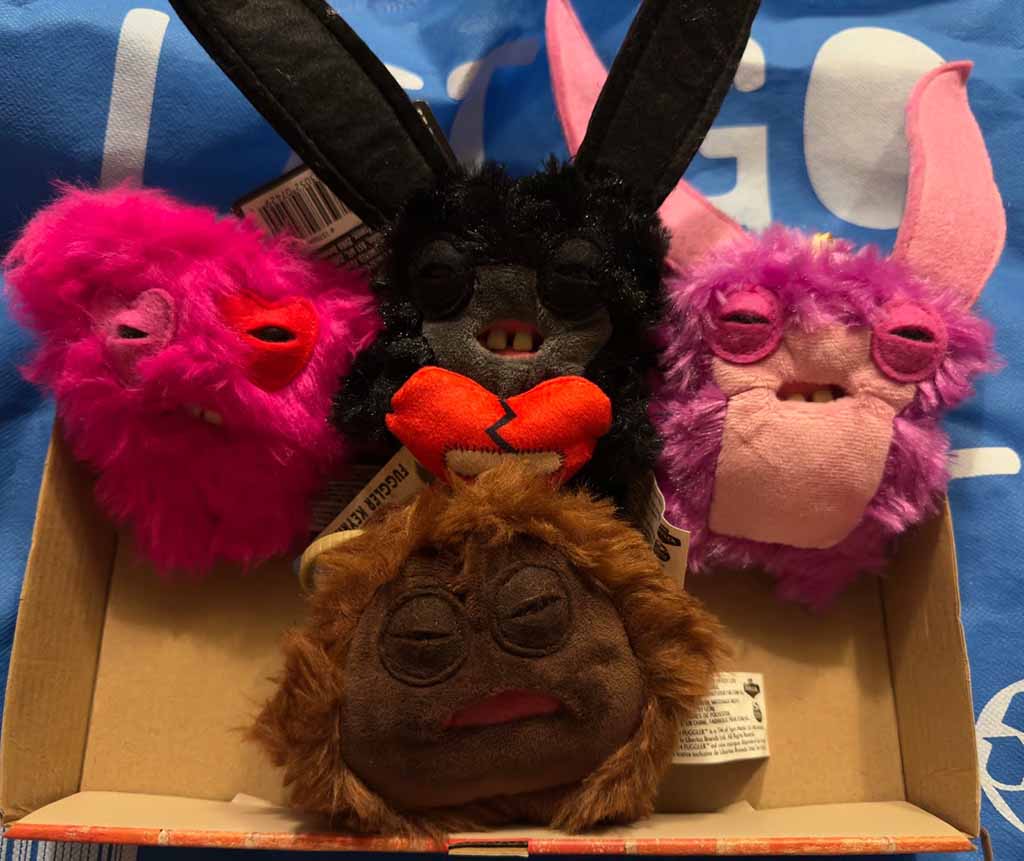 Collection of quirky Fuggler plush toys in bright colors—pink, black, purple, and brown—displayed in an open box, each featuring their signature silly teeth and expressive faces.