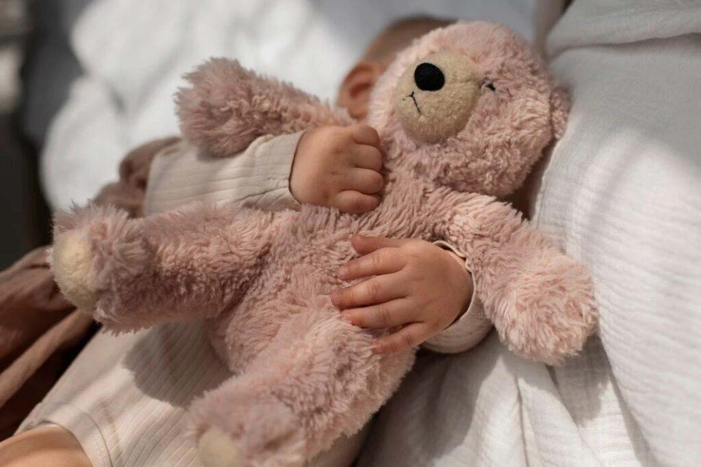 Close-up of a newborn baby gently holding a soft plush teddy bear while resting on bedding, highlighting emotional comfort, tactile softness, and infant-friendly plush toy design without implying unsupervised sleep use.