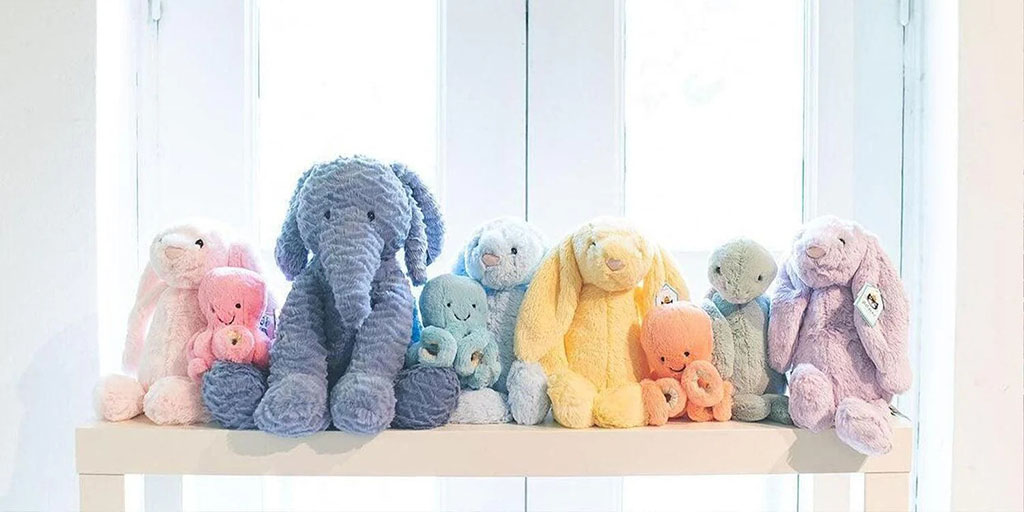 Row of pastel-colored plush animals, including bunnies, elephants, and octopus toys, neatly arranged on a white shelf in front of bright window light.