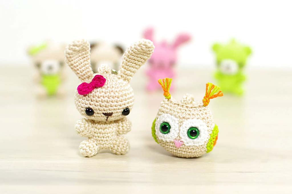 Small handmade crochet bunny with a pink bow and a crochet owl with green eyes placed on a light wooden surface, with colorful blurred amigurumi figures in the background.