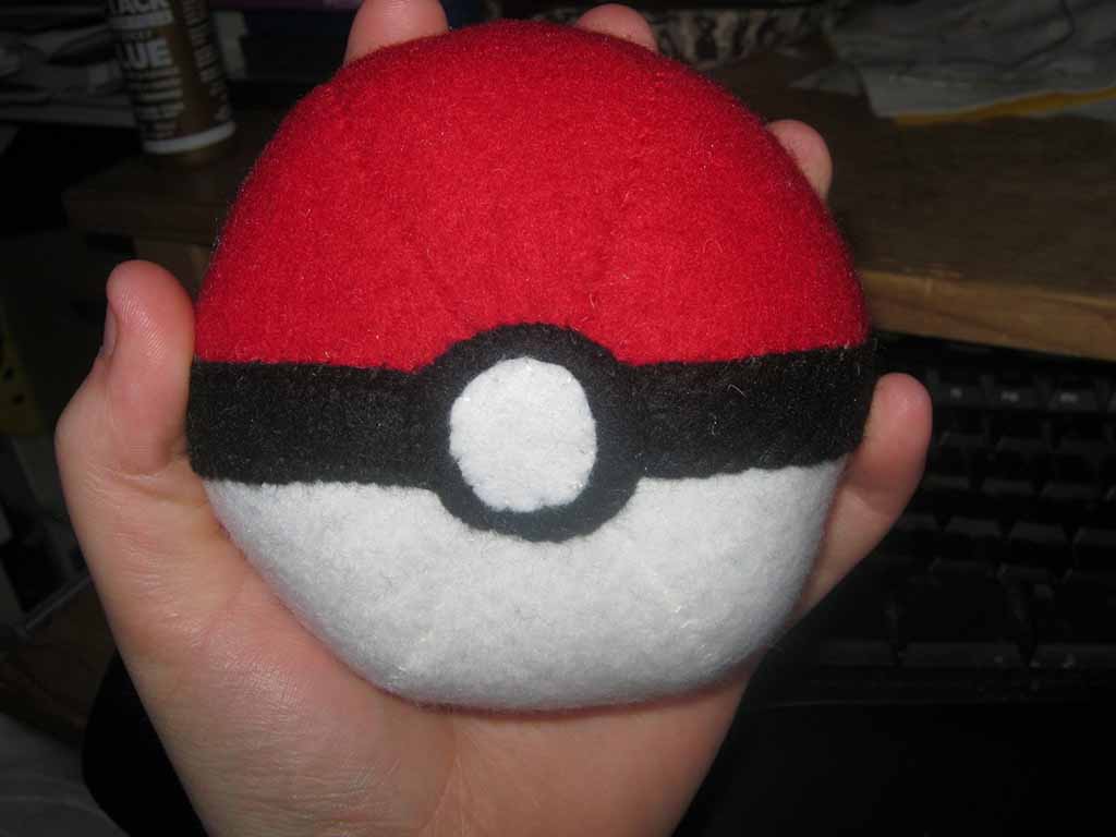 Hand holding a round red, white, and black plush ball with a central circular detail, resembling a Poké Ball design, showing soft fleece texture and handmade stitching.