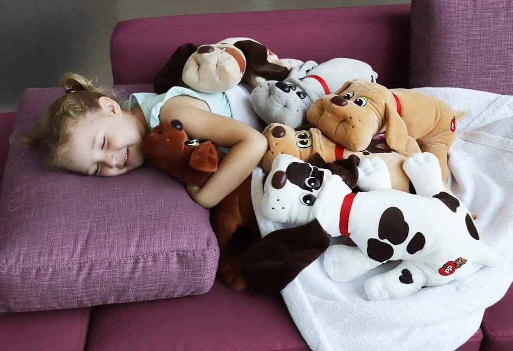 A young girl happily cuddles a pile of soft plush dog toys while resting on a purple sofa, surrounded by multiple stuffed puppies in various colors and breeds.