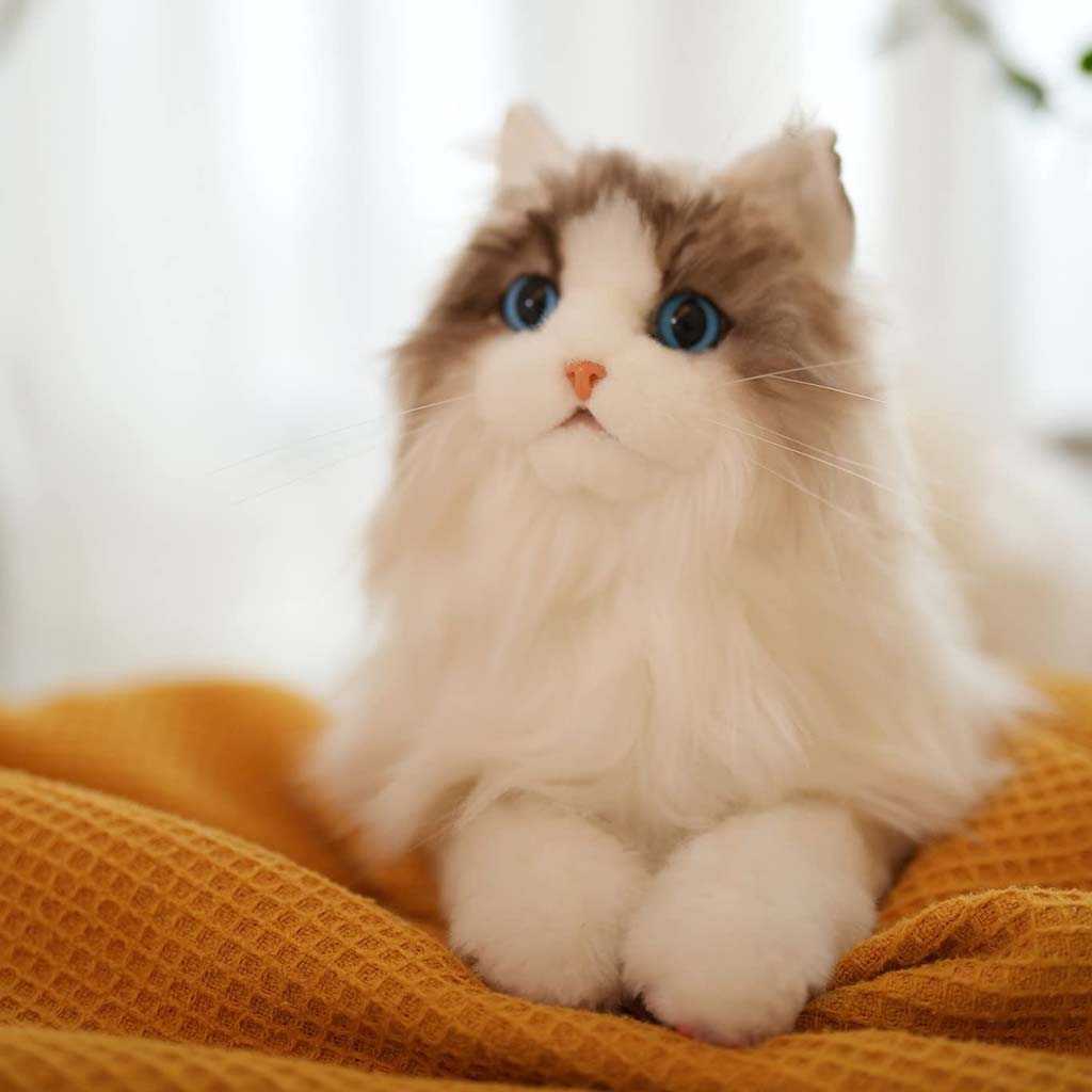 A realistic long-haired cat plush with soft white and gray fur, bright blue eyes, and a pink nose, resting on a textured mustard blanket in soft natural lighting.