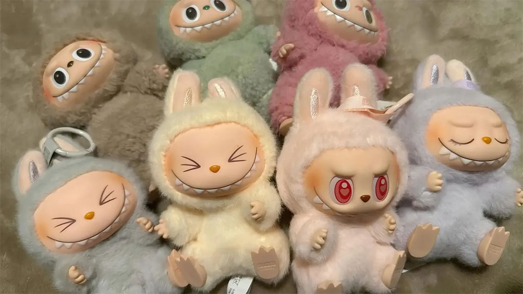 A pastel-colored collection of Labubu plush dolls arranged on a soft surface, featuring multiple expressions and outfits, including smiling, sleepy, and red-eyed variants in shades of beige, pink, grey, and green.