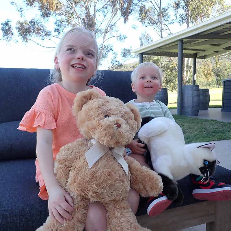 Two young children sit outdoors on a bench, smiling and holding plush toys including a large teddy bear and a soft cat doll, illustrating how stuffed animals support comfort, play, and emotional bonding.