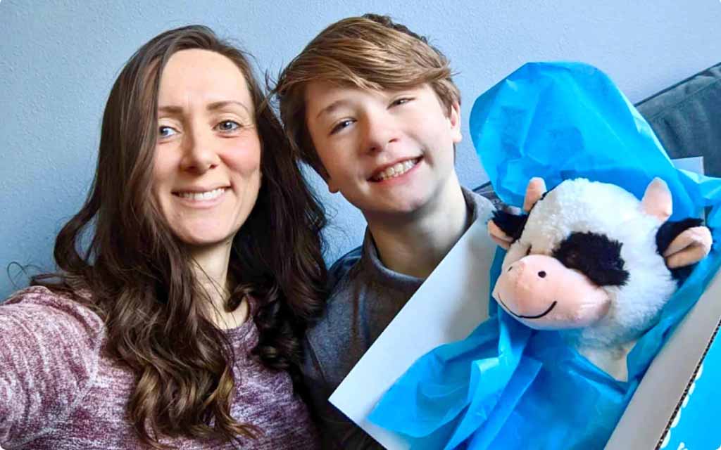 A woman and a boy smiling while holding a gift box containing a soft cow plush toy wrapped in blue tissue paper.