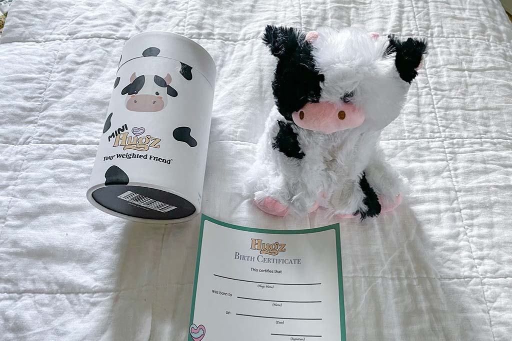 A mini weighted cow plush toy displayed on a white quilt alongside its branded cylindrical packaging and a printed birth certificate, highlighting a compact weighted stuffed animal gift set designed for comfort, emotional support, and calming sensory use, with soft textures and premium presentation suitable for retail or gifting.