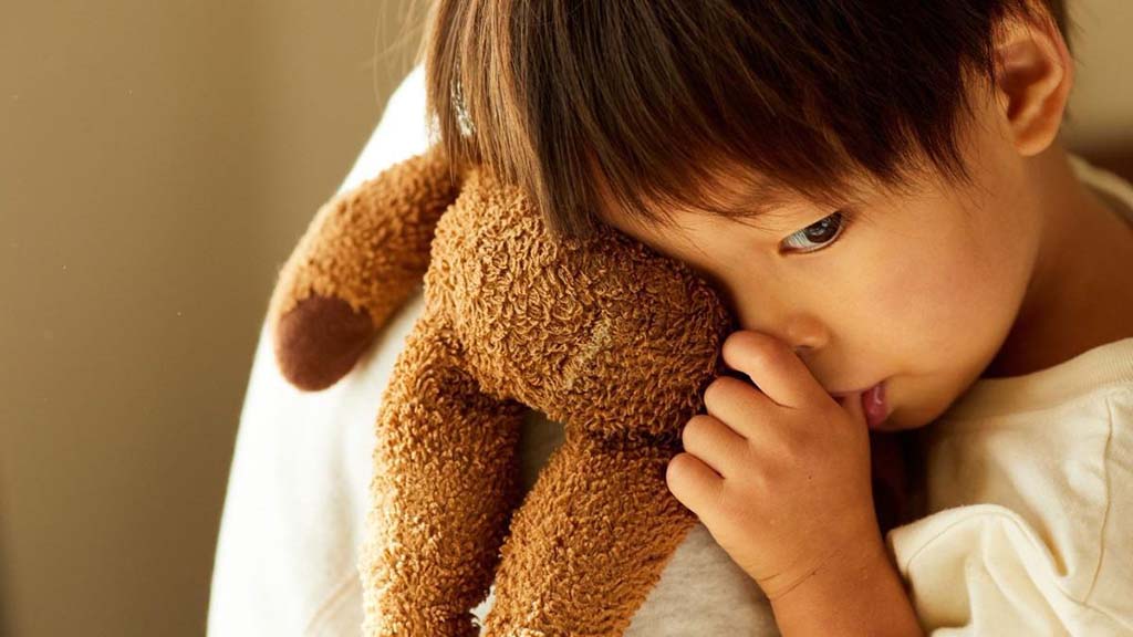 A young child with short dark hair holds a stuffed brown bear closely while sucking their thumb. The child rests their head against the toy, creating an intimate and comforting moment. The soft, neutral background highlights the child's affection for the plush toy.