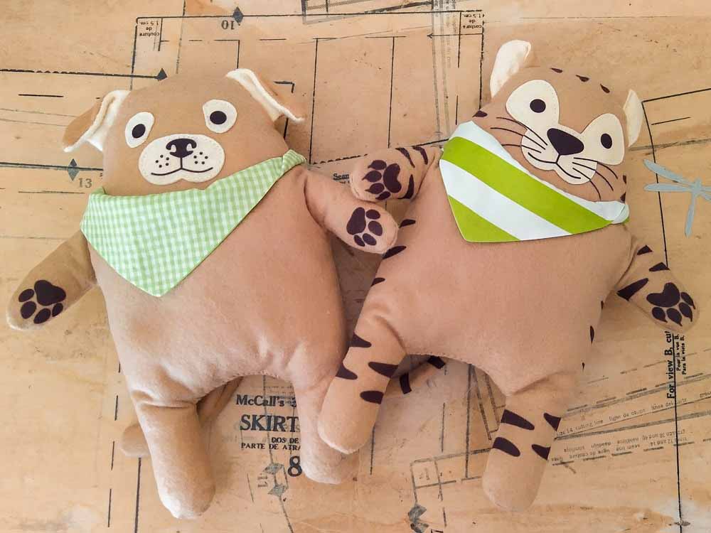 Two handmade plush toys—a dog and a tiger—lying on a sewing pattern background. Both toys have soft tan bodies, printed facial features, paw details, and green bandanas (one gingham, one striped). Their rounded shapes and simple limbs give them a cute, friendly look.