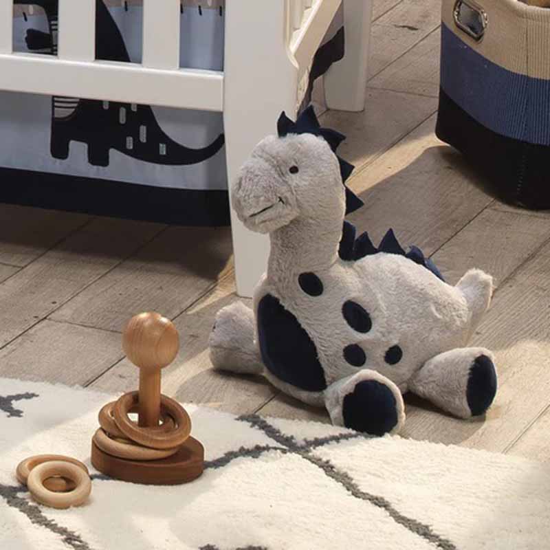 Soft dinosaur plush toy placed on a nursery floor beside a wooden ring-stacking toy and crib, featuring neutral gray and black tones, rounded shapes, embroidered facial details, and child-safe materials, suitable for baby rooms, infant gifting, and OEM or wholesale plush toy manufacturing.