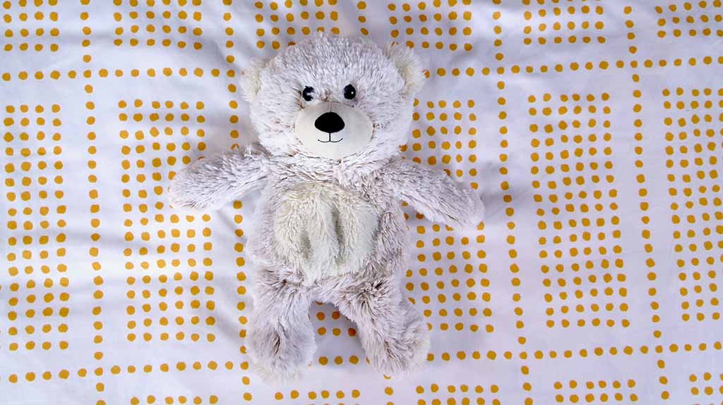A light grey plush teddy bear lying on a white bedsheet with a yellow dotted pattern.