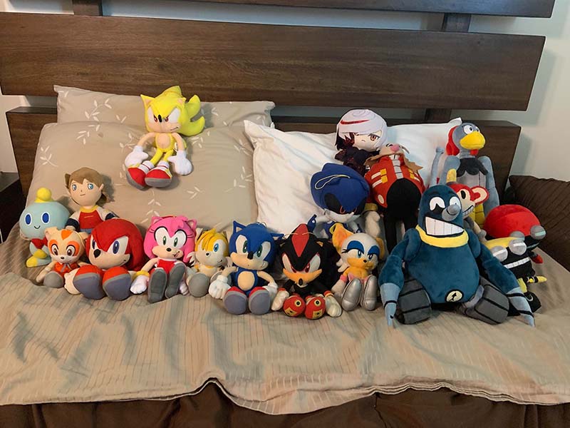 Collection of licensed character plush toys arranged on a bed, featuring multiple popular game and cartoon characters in soft stuffed toy form, demonstrating character-based plush design, licensed plush manufacturing, and detailed OEM plush toy production for branded merchandise lines.